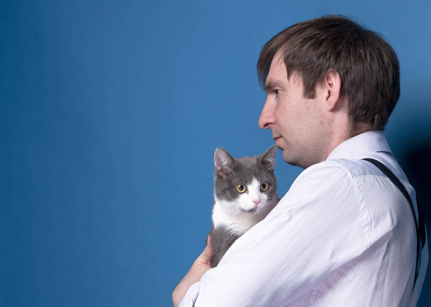 side view of man in pink shirt holding grey cat on blue background with copy space - Fotoğraf, Görsel