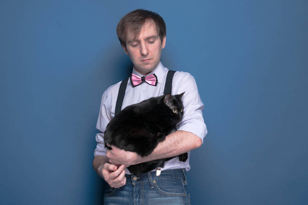 confident handsome man in shirt, suspender and pink bow tie holding and looking to cute black cat on blue background with copy space - Foto, immagini