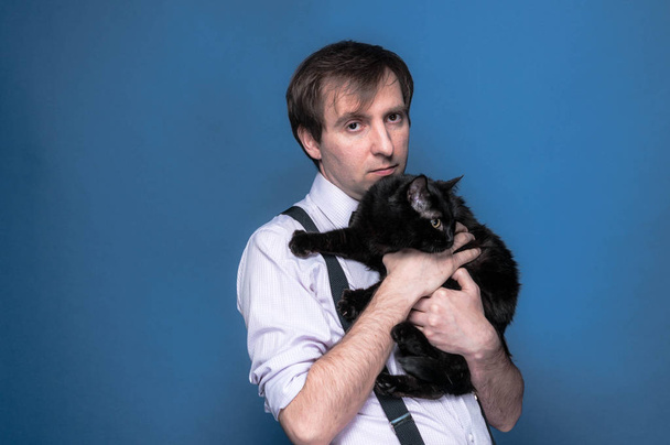 handsome man in shirt and suspender holding and hugging black funny cat on blue background - Photo, image