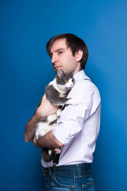 side view of handsome man in shirt and black suspenders, holding and stroking grey cat in golden crown on blue background with copy space - Valokuva, kuva