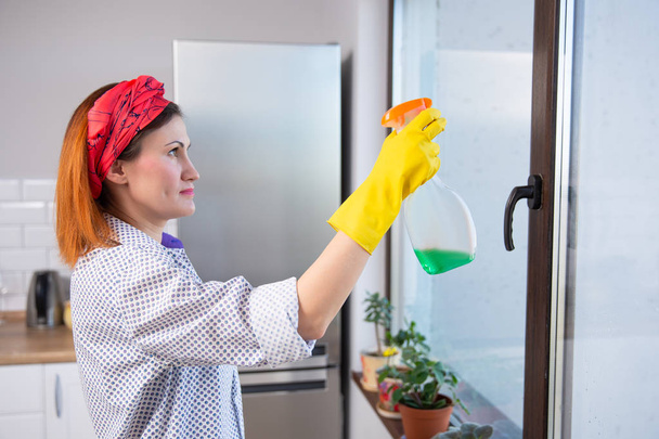 people, housework and housekeeping concept - woman in gloves cleaning window with cleanser spray at home - Photo, Image