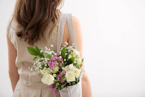 Young woman with bouquet of flowers in eco bag on light background - Photo, Image