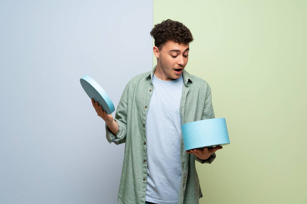 Jonge man over blauwe en groene achtergrond die een gift in handen houdt - Foto, afbeelding