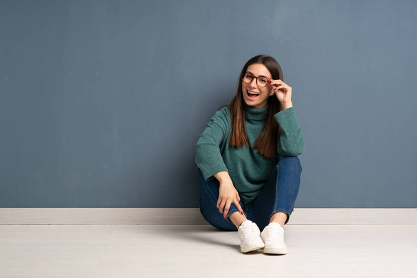 Νεαρή γυναίκα κάθεται στο πάτωμα με γυαλιά και έκπληκτος - Φωτογραφία, εικόνα