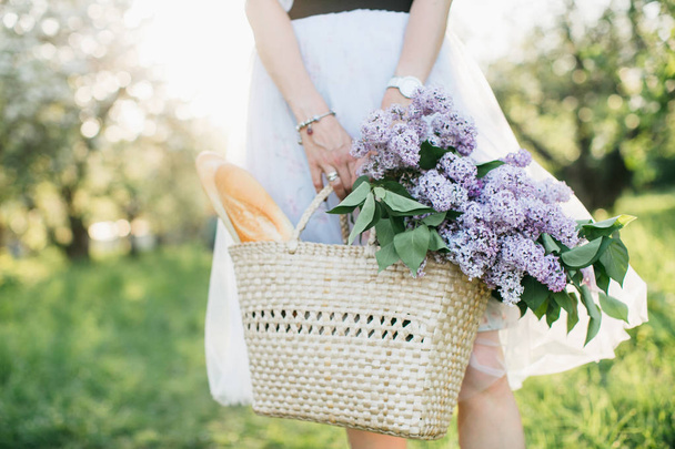 Basket with bouquet of lilacs and baguette in woman hands on bac - Fotó, kép