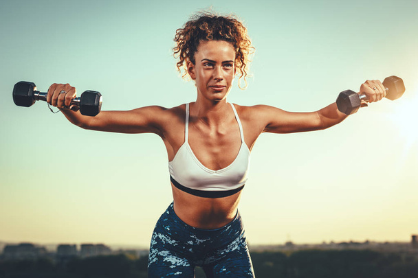 Young fitness woman doing workout with dumbbells on sunset background - Photo, Image