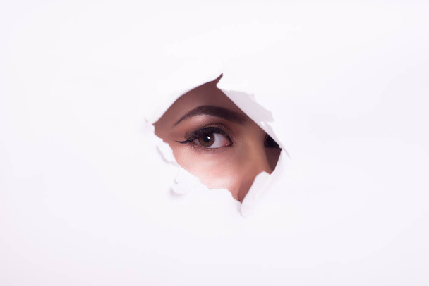 eyes of a young curious woman peeking through a hole torn in white paper poster. with beautiful make-up. - Fotografie, Obrázek