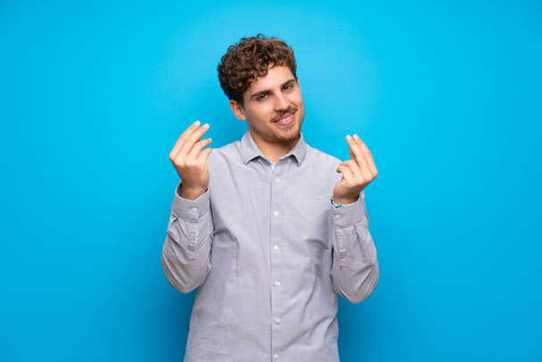 Blonde man over blue wall making money gesture - Photo, Image