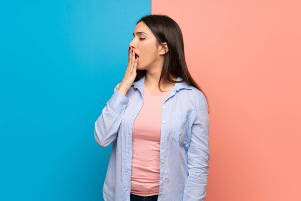 Young woman over pink and blue wall yawning and covering wide open mouth with hand - Foto, imagen