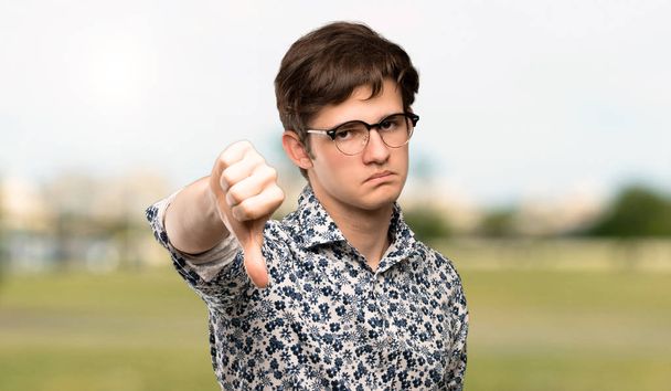 Homme adolescent avec chemise à fleurs et lunettes montrant pouce vers le bas avec expression négative à l'extérieur
 - Photo, image