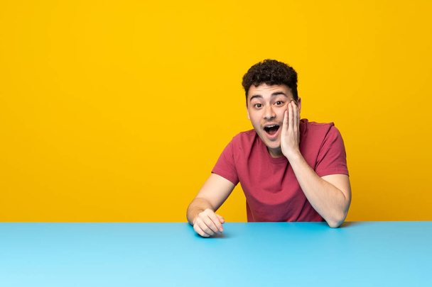 Giovane uomo con parete colorata e tavolo con sorpresa e espressione facciale scioccata
 - Foto, immagini