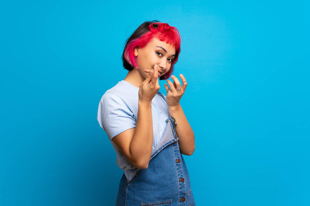 Fiatal nő, rózsaszín haj kék falon, így pénzt gesztus - Fotó, kép