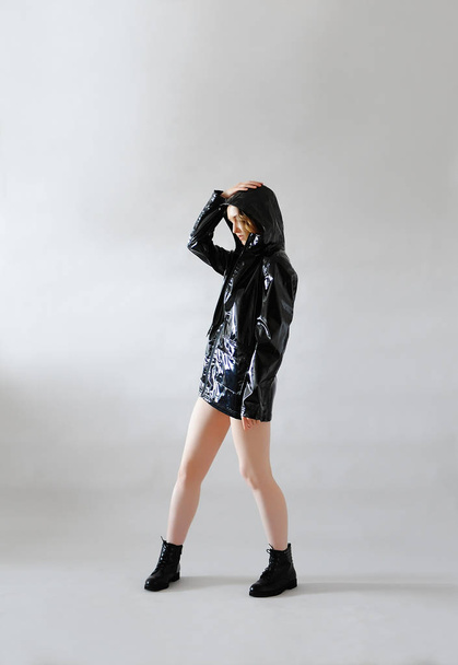 Jeune fille élégante en veste de pluie laquée noire imperméable avec capuche et chaussures basses. Fond clair. Tenue tendance
 - Photo, image