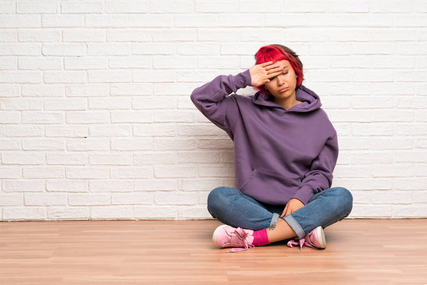 Young woman with pink hair sitting on the floor with tired and sick expression - 写真・画像