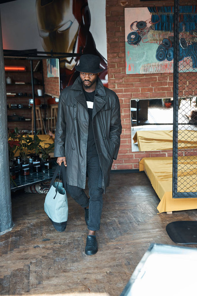 Fashionable young man in black waxed trench coat, bucket hat and with blue bag, walking in loft interior room with painting and chain link.  - Foto, imagen