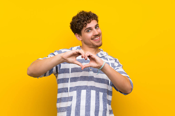 Homme blond sur le mur jaune faisant symbole de coeur par les mains
 - Photo, image