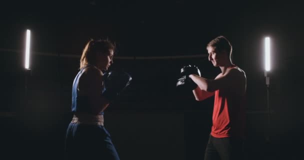 schöne brünette Boxerin, die in einem dunklen Raum mit einem Trainer Schläge auf die Pfoten trainiert - Filmmaterial, Video