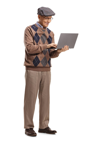 Full length portrait of a senior man standing and using a laptop isolated on white background - Фото, изображение