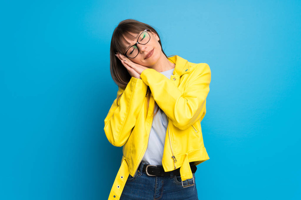 Jonge vrouw met gele jas op blauwe achtergrond maken slaap gebaar in dorable expressie - Foto, afbeelding