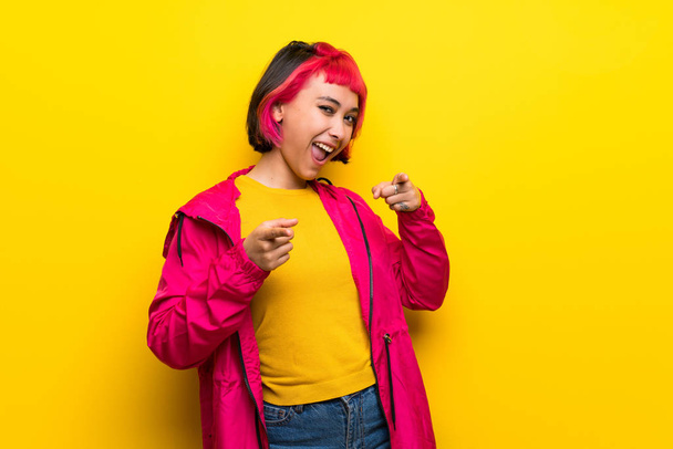Mujer joven con el pelo rosa sobre la pared amarilla señalando hacia el frente y sonriendo
 - Foto, imagen