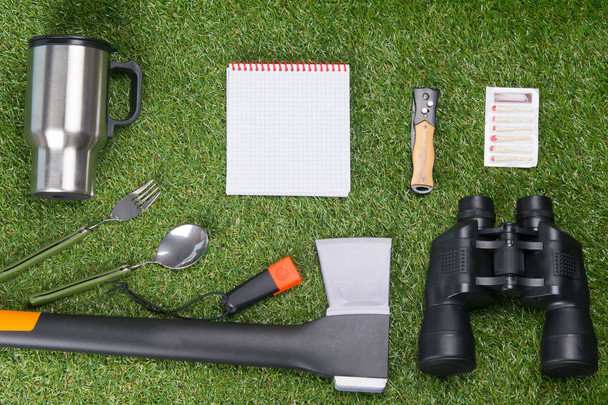 against the background of a green lawn, a set of objects of the traveler, an ax, binoculars, tourist matches, a folding knife, a fork, a spoon, a flashlight, there is a place for an inscription - Фото, изображение