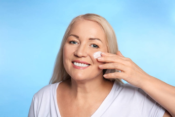 Mature woman applying facial cream on color background - Photo, Image