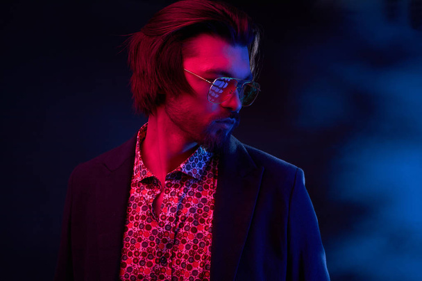 Neon light portrait of handsome male model with dark medium length hair, mustaches and beard in sunglasses, weared in dark jacket and floral shirt. - Фото, зображення
