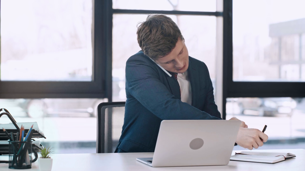homem bonito digitando no laptop, olhando para o smartphone, falando, sorrindo e escrevendo no notebook no escritório moderno
  - Filmagem, Vídeo