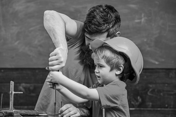 Father, parent with beard teaching little son to use tool screwdriver. Teamwork in workshop concept. Boy, child busy in protective helmet learning to use screwdriver with dad - Photo, Image