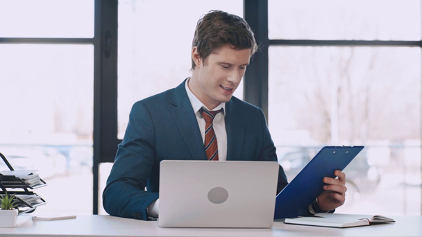 handsome businessman holding clipboard, looking at contract while holding pen and using laptop and celebrating triumph in modern office  - Footage, Video