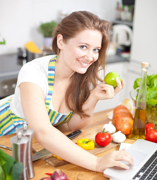 young woman in apron smiling at camera and using laptop in kitchen - 写真・画像