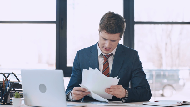 αναστατωμένος επιχειρηματίας κοιτάζοντας τις συμβάσεις, ενώ καθισμένος κοντά σε φορητό υπολογιστή και ρίχνοντας χαρτιά στον αέρα στο σύγχρονο γραφείο  - Πλάνα, βίντεο