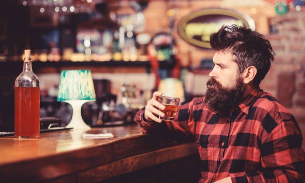 Guy spend leisure in bar, defocused background. Hipster with beard ordered full bottle of alcohol. Relaxation concept. Man drinks whiskey or cognac. Man with strict face sits near bar counter - Photo, image