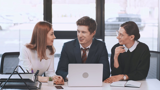 knappe zakenman zit met aantrekkelijke medewerkers en kijken naar laptop, praten en glimlachen in de buurt van vrouw schrijven in notebook  - Video