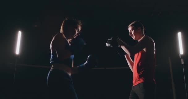 Beautiful brunette boxer working out blows to the paws with a trainer in a dark room - Footage, Video