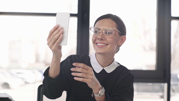 cheerful woman in glasses holding smartphone while having video chat and waving hand in office  - Filmagem, Vídeo