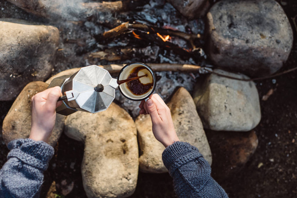 View from above of traveler woman pours itself hot beverage in mountains near to bonfire. Female sitting and holding a mug of coffee after hiking. Travel, people and healthy lifestyle concept - Photo, image