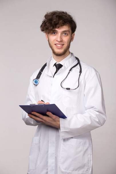 Portrait of smiling confident young smiling medical doctor with sthetoscope and clipboard over light gray background - Photo, Image