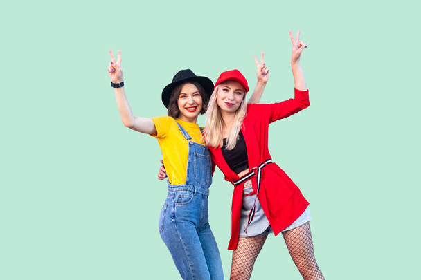 two beautiful happy fashionable hipster women standing and showing v sings while looking at camera on green background. - Fotó, kép