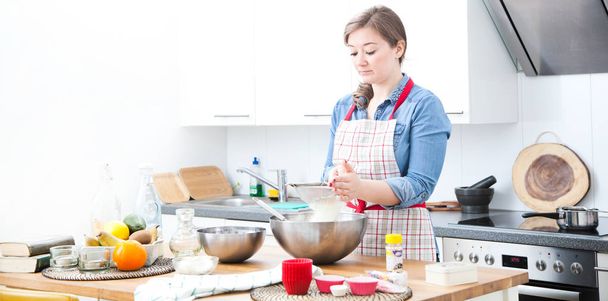 beautiful young woman in apron preparing dough for cupcakes in kitchen - Photo, image