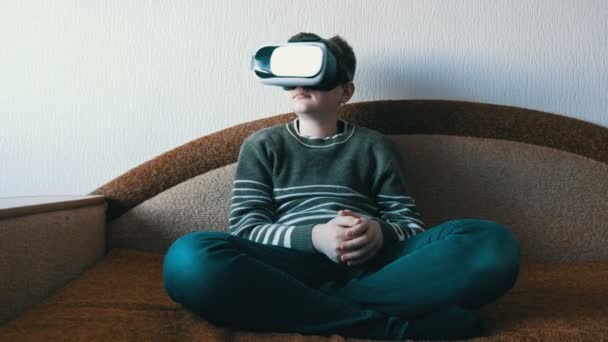 Tini fiú fehér pohár virtuális valóság vagy VR a fejét ül a kanapén a szobában és a játék. - Felvétel, videó