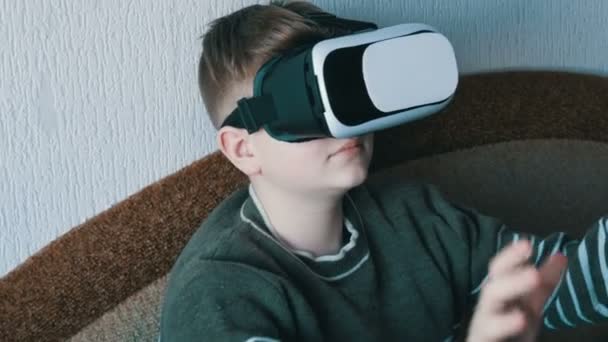 Adolescente niño en gafas blancas de realidad virtual o vr en su cabeza sentado en el sofá en la habitación y jugando
. - Imágenes, Vídeo