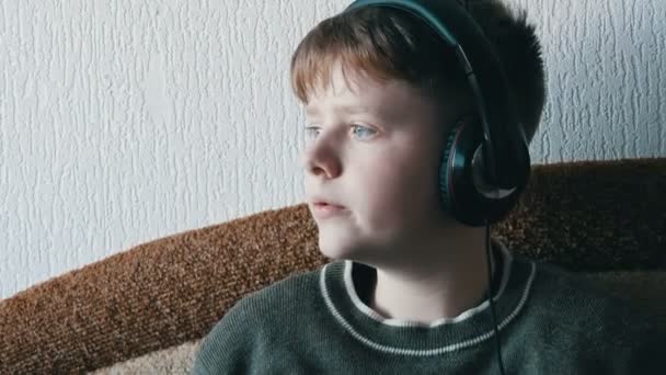 Хлопчик з великими навушниками на голові слухає музику на смартфоні, сидячи на коричневому дивані
. - Кадри, відео