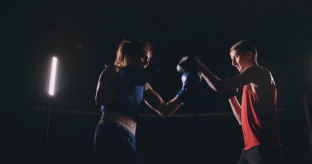 Beautiful brunette boxer working out blows to the paws with a trainer in a dark room - Footage, Video
