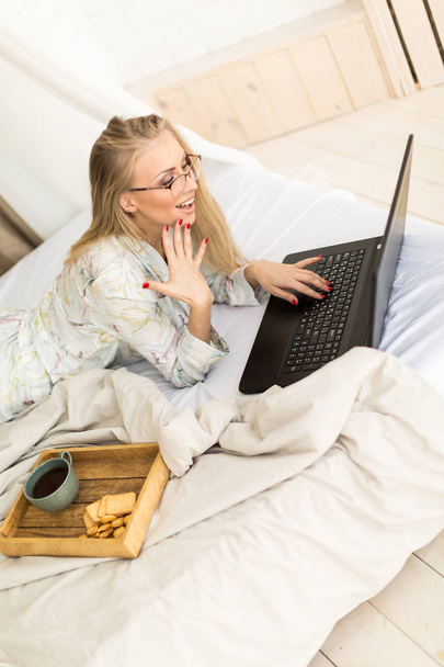 Young smiling woman in glasses with cup of coffee and laptop lying on the bed. Vintage brick wall on background. - Photo, image