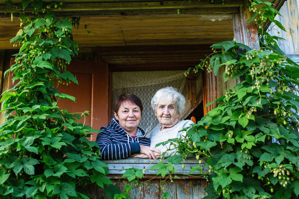 Ηλικιωμένη γυναίκα και κόρη ενηλίκων κοιτάζοντας έξω από το σπίτι του χωριού. - Φωτογραφία, εικόνα