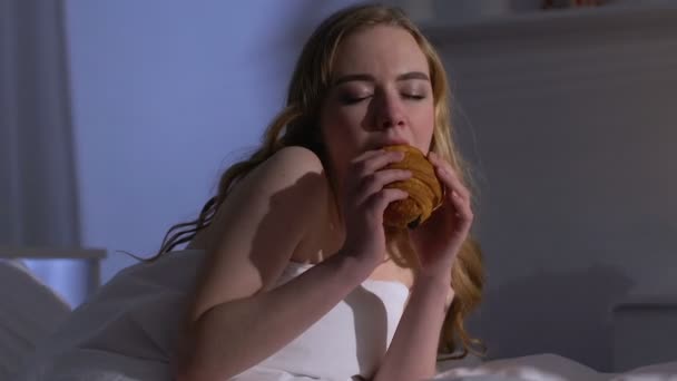 Woman eating croissant in bed at night, enjoying sweets after diet, bulimia - 映像、動画