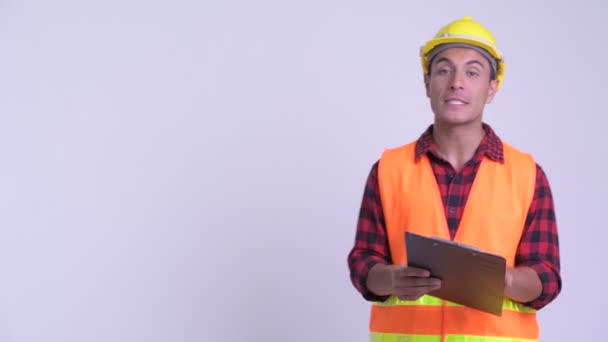 Young happy Hispanic man construction worker presenting with blackboard and clipboard - Кадры, видео