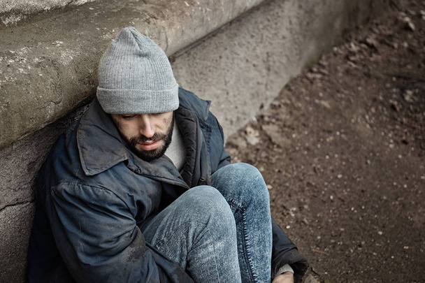 Poor homeless man sitting near wall on street. Space for text - Zdjęcie, obraz
