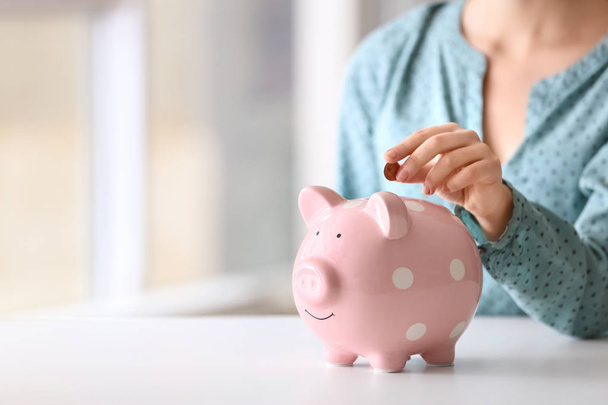 Woman putting coin into piggy bank at table indoors, closeup. Space for text - Photo, Image